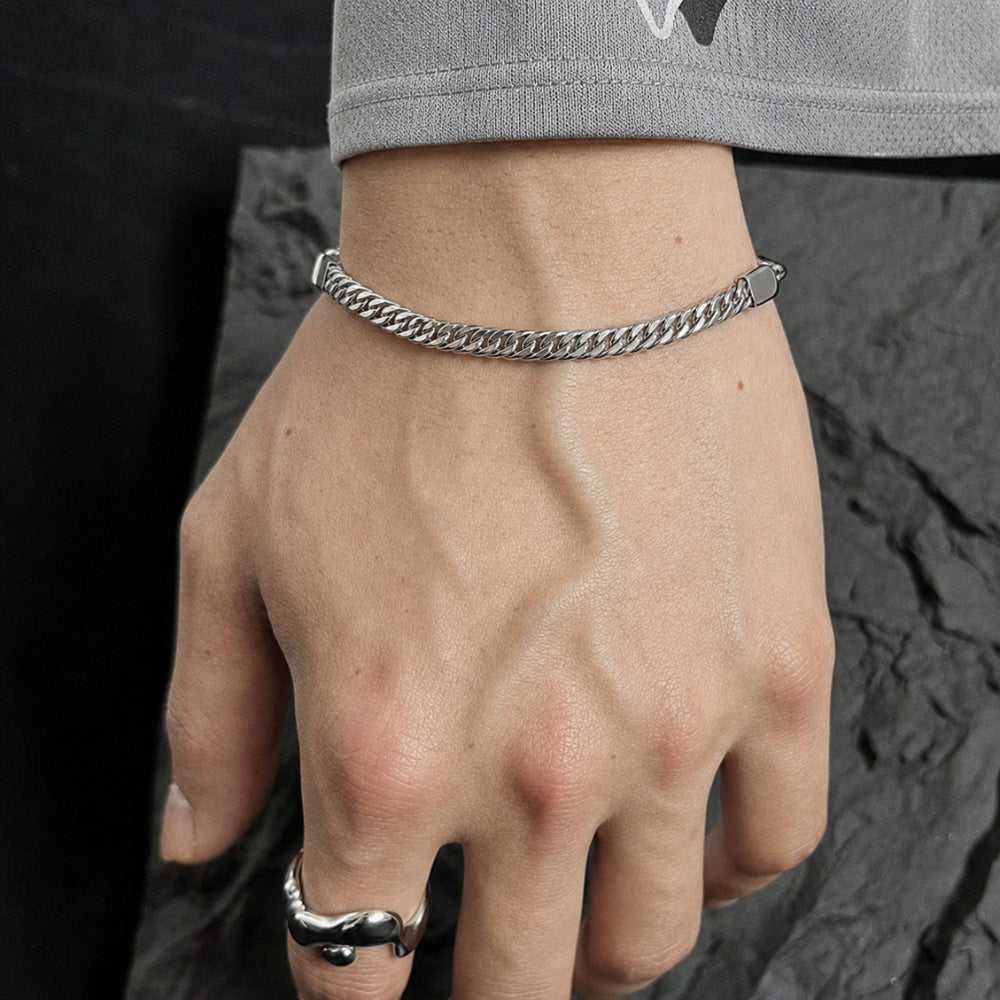 A close-up of a person's hand wearing a silver chain bracelet and a silver ring, with a gray sleeve visible at the top against a dark textured background.