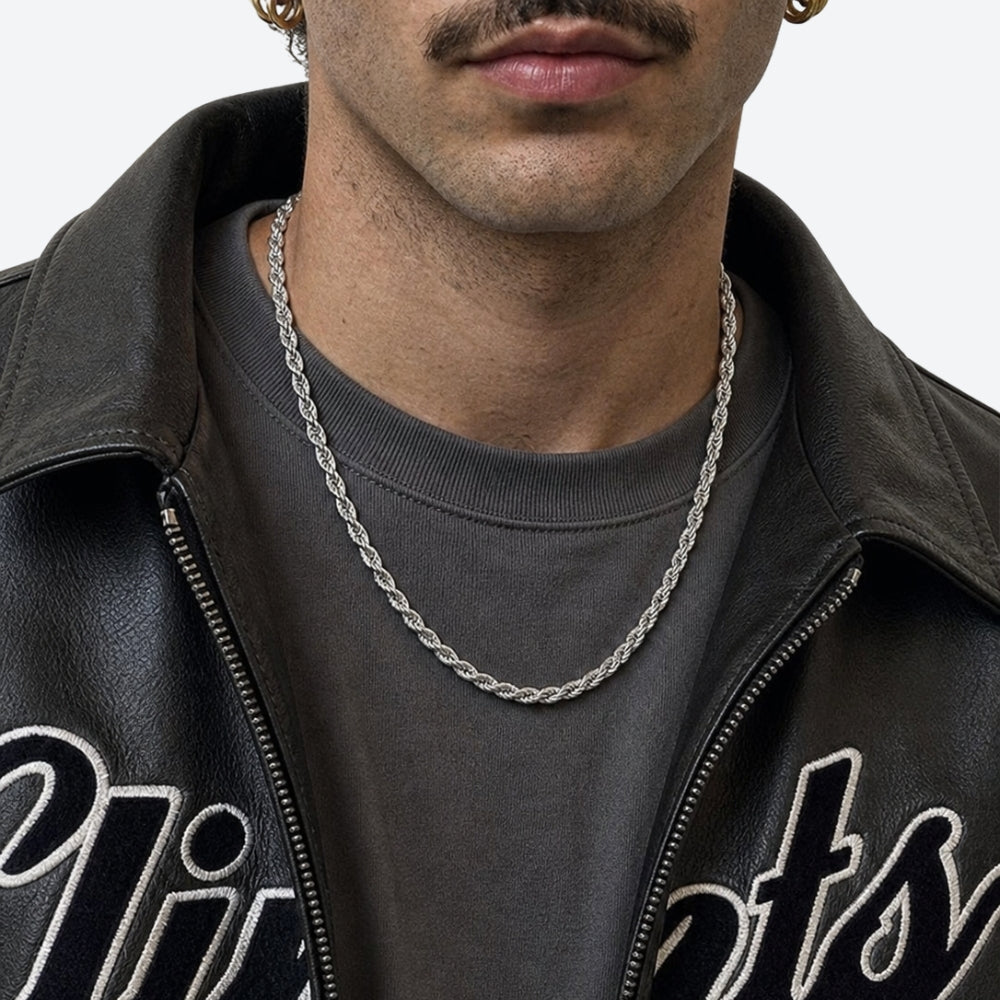 A close-up of a man wearing a silver chain necklace over a dark gray t-shirt and a black leather jacket.
