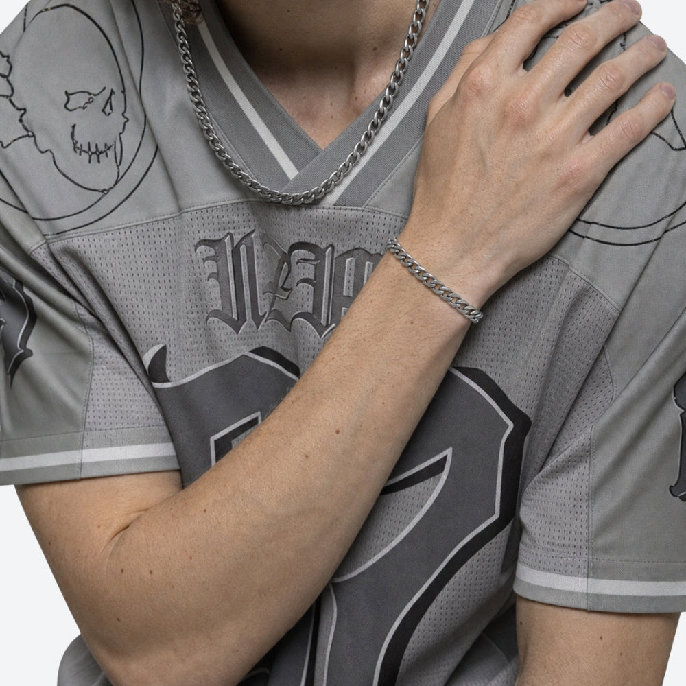 Close-up of a person wearing a gray and black graphic jersey, a silver chain necklace, and a silver chain bracelet, with one hand resting on the other shoulder.