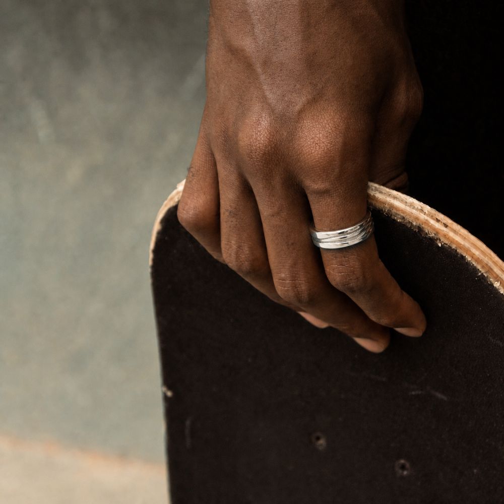 A close-up of a person's hand wearing a textured silver band ring, holding the edge of a black skateboard deck.