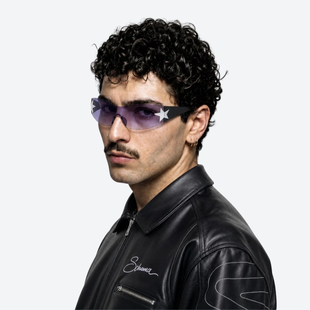 A man with curly black hair and a mustache wearing purple-tinted sunglasses with a white star detail on the frame, paired with a black leather jacket, shown against a light gray background.