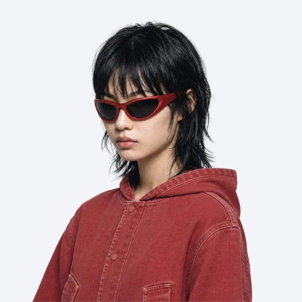 A model with short black hair wearing red cat-eye sunglasses and a matching red hooded jacket, photographed against a light gray background.