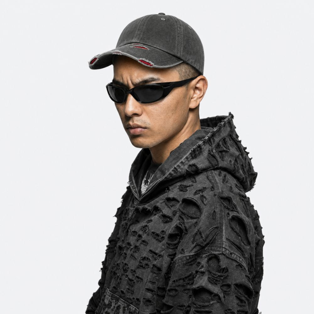 A man in profile wearing a dark gray distressed baseball cap, black sunglasses, and a heavily textured black hooded jacket against a plain light gray background.