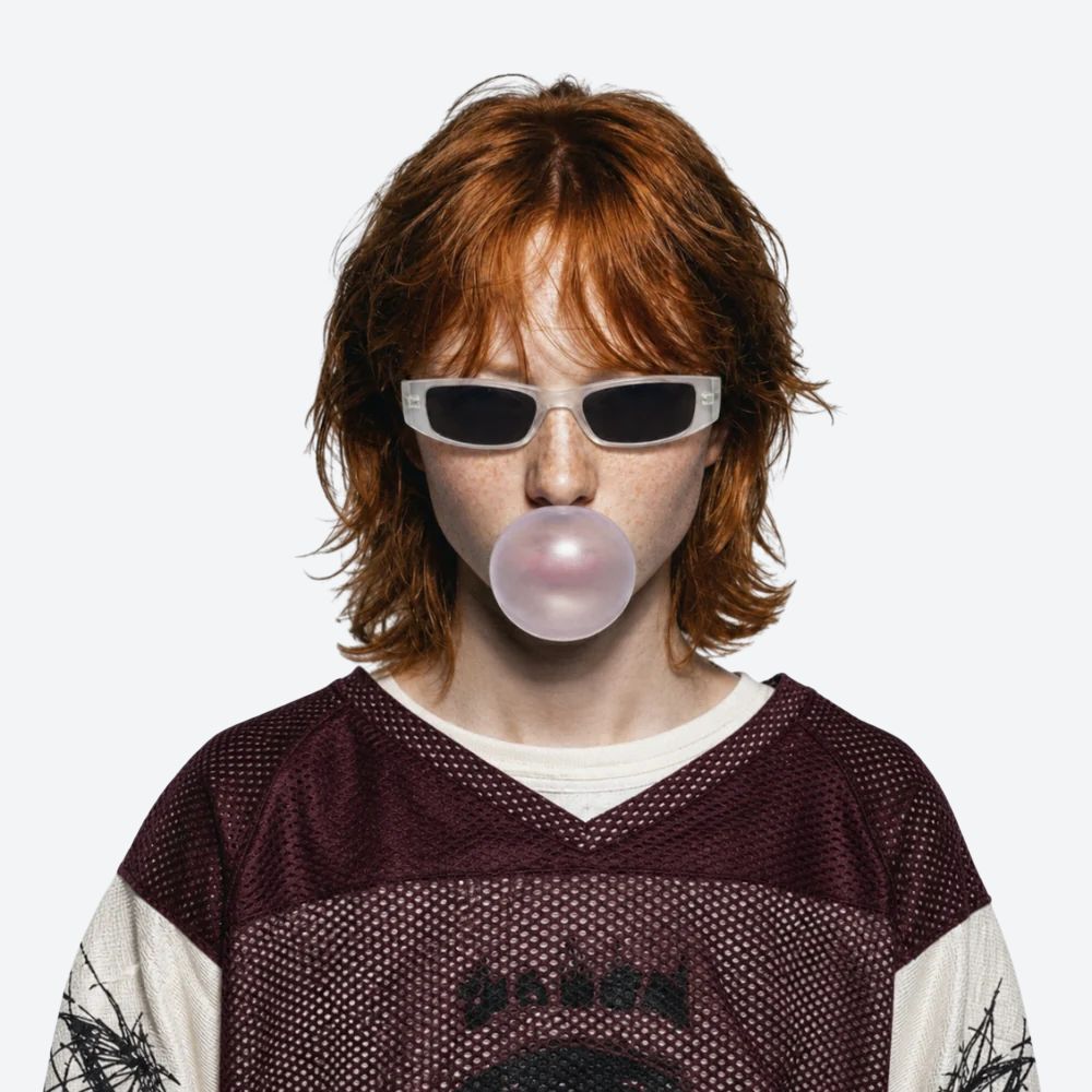 A person with red hair wearing white-framed dark sunglasses and a burgundy-and-white mesh shirt, blowing a pink bubble gum bubble against a plain light gray background.