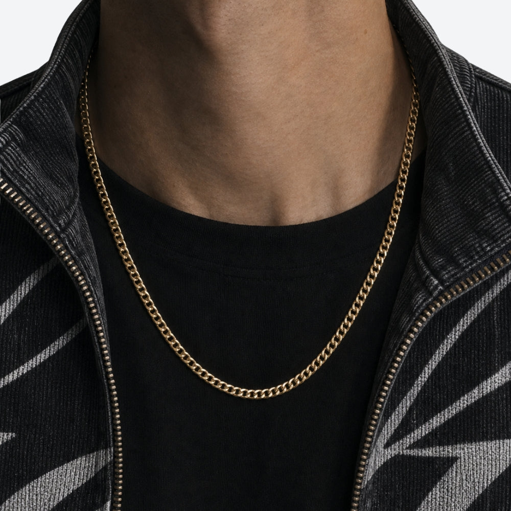 A close-up of a person's neck and chest wearing a gold chain necklace over a black t-shirt and a dark patterned zip-up jacket.