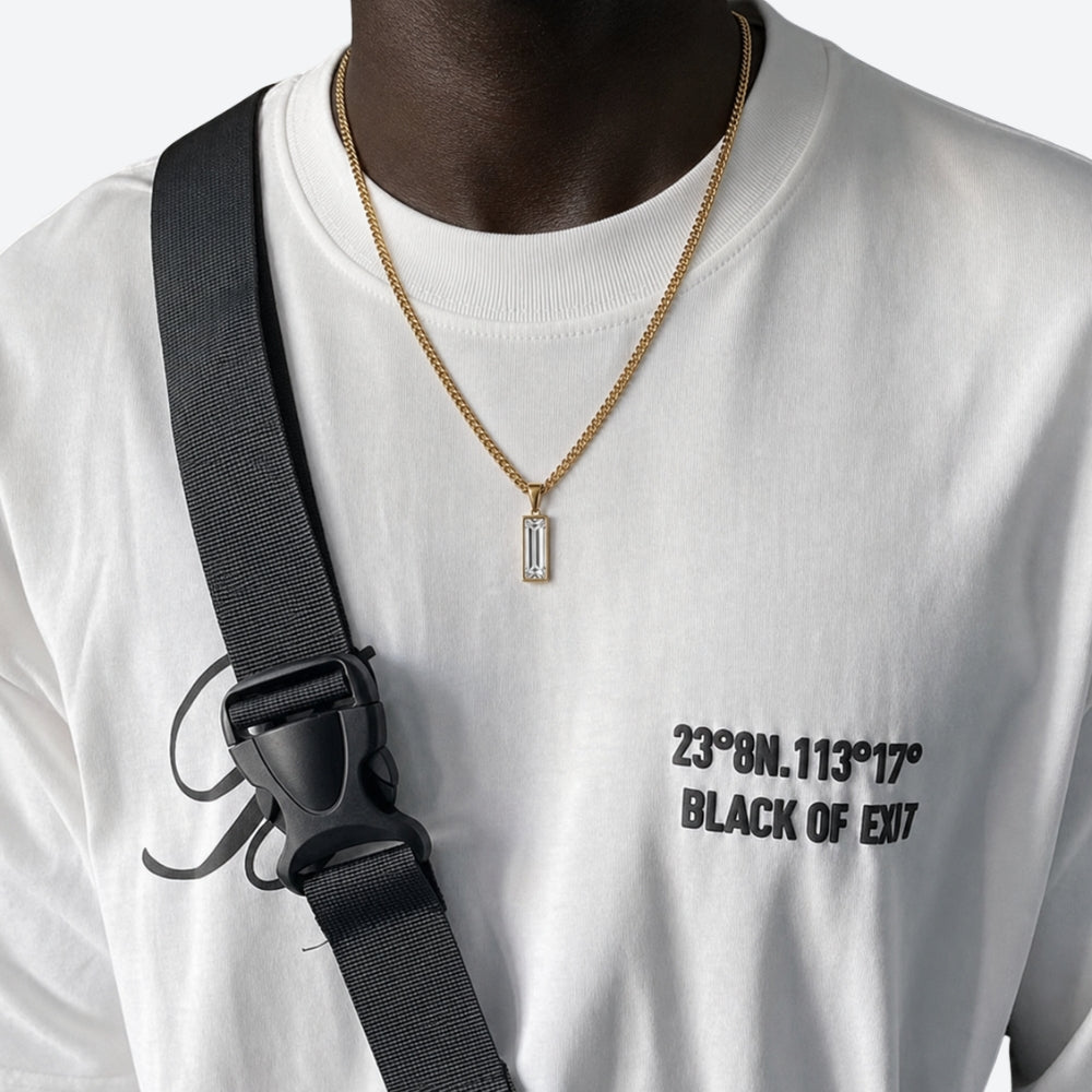 A close-up of a person wearing a gold chain necklace with a rectangular baguette pendant, styled over a white graphic t-shirt with a black crossbody strap.
